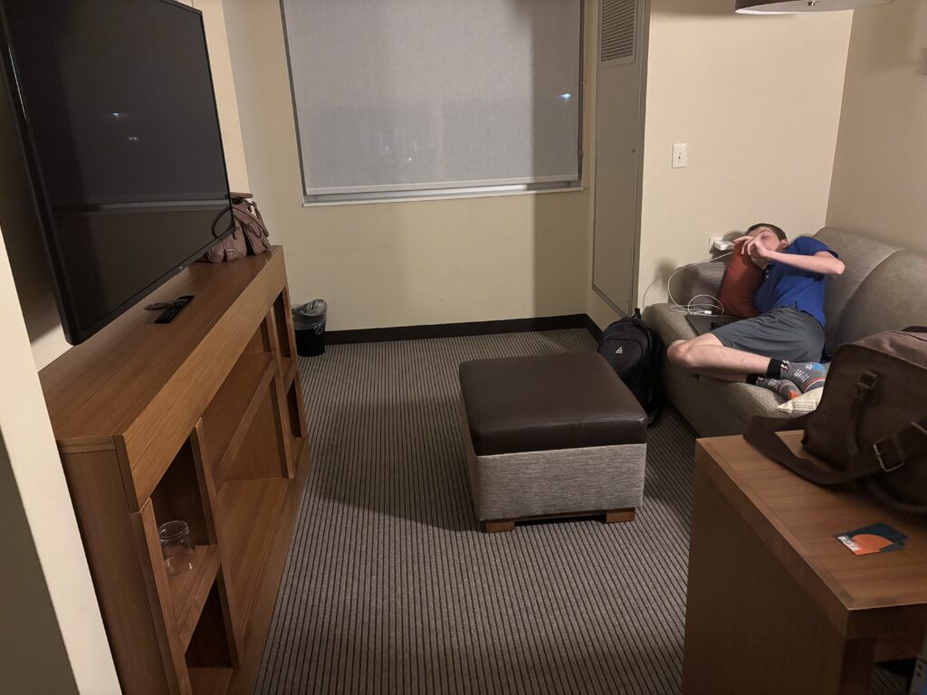 A person is lying on a couch in a hotel room. The room has a large flat-screen TV mounted on a wooden stand, a small ottoman, and a window with a closed blind. There is a backpack on the floor next to the couch and a brown bag on a table. The person is wearing a blue shirt and gray shorts.