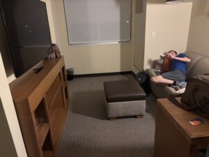 A person is lying on a couch in a hotel room. The room has a large flat-screen TV mounted on a wooden stand, a small ottoman, and a window with a closed blind. There is a backpack on the floor next to the couch and a brown bag on a table. The person is wearing a blue shirt and gray shorts.