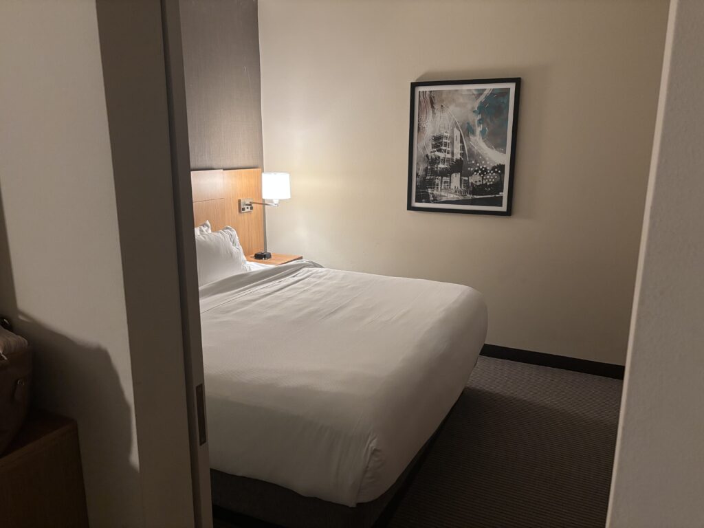 The image shows a hotel room with a neatly made bed featuring white linens. A wooden headboard is visible, and there is a bedside table with a lamp on it. A framed picture hangs on the wall above the bed. The room has a neutral color scheme with beige walls and carpet.