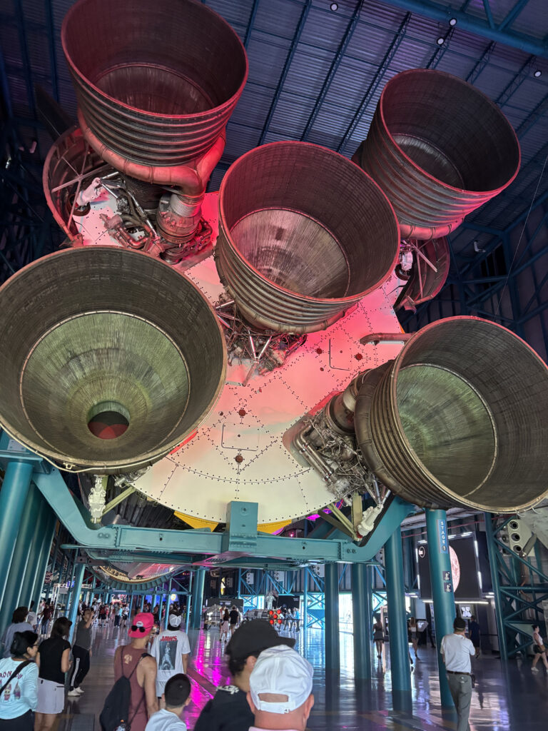 The image shows the underside of a large rocket with five massive engine nozzles. The rocket is displayed indoors, and there are people walking underneath it, giving a sense of scale. The area is well-lit, with some red and purple lighting highlighting the engines. The structure is supported by blue beams, and the ceiling is visible above.