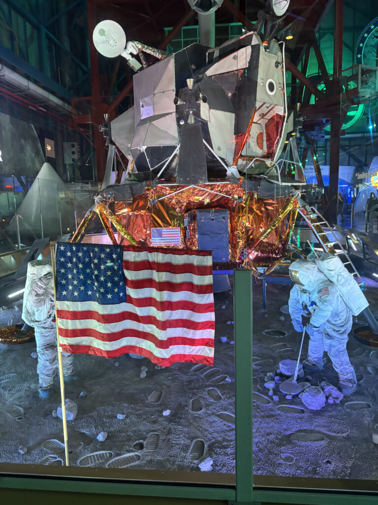 A display of a lunar module with two astronaut figures in spacesuits on a simulated moon surface. One astronaut is planting an American flag, while the other is examining rocks. The scene includes footprints and rocks on the ground, with the lunar module in the background.