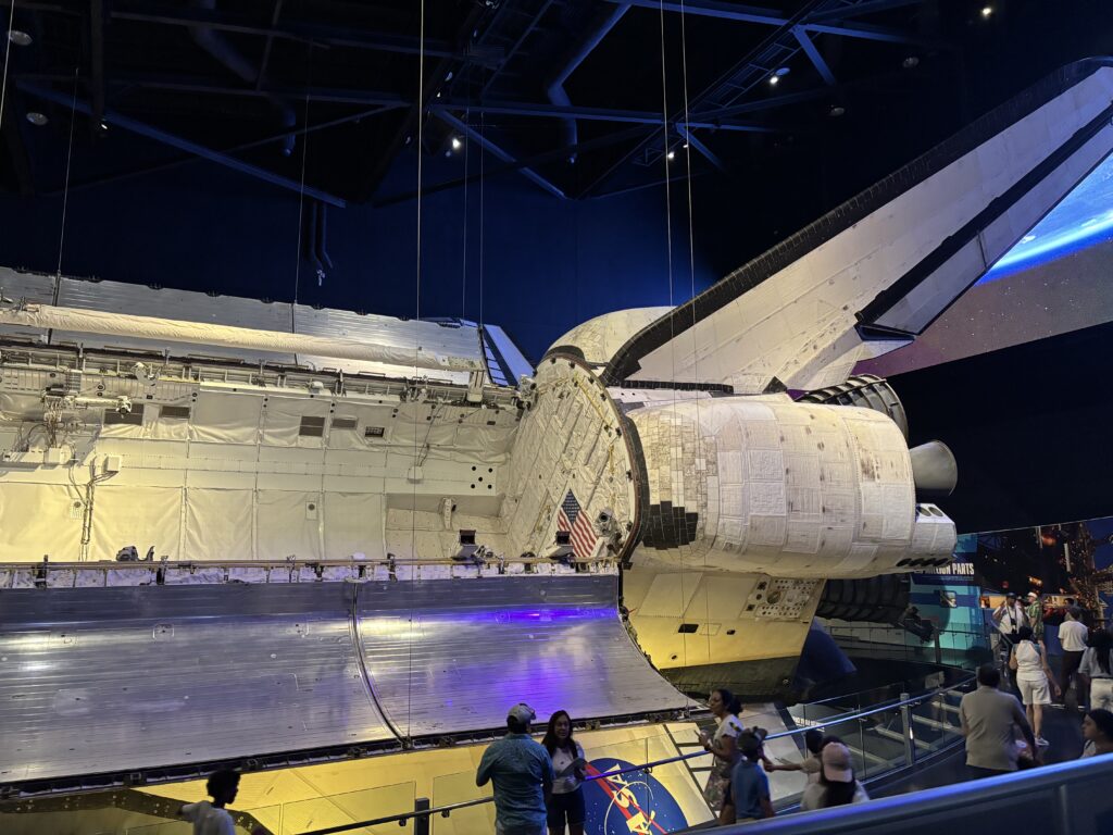 The image shows a space shuttle displayed in a museum setting. The shuttle is suspended, allowing visitors to view it from below. The shuttle's cargo bay doors are open, and its large tail fin and engines are visible. People are walking around and observing the exhibit. The lighting is dim, with spotlights highlighting the shuttle.