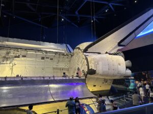 The image shows a space shuttle displayed in a museum setting. The shuttle is suspended, allowing visitors to view it from below. The shuttle's cargo bay doors are open, and its large tail fin and engines are visible. People are walking around and observing the exhibit. The lighting is dim, with spotlights highlighting the shuttle.