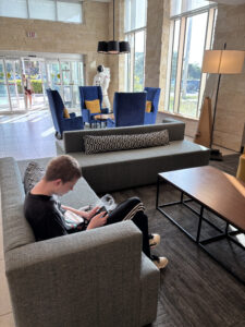 A person is sitting on a gray sofa in a modern lobby, looking at a phone. The room has large windows, blue chairs with yellow cushions, and a wooden coffee table. A life-sized astronaut figure is near the entrance, and a person is entering through the glass doors. The space is well-lit with natural light.