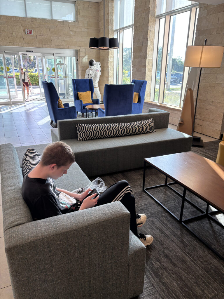 A person is sitting on a gray sofa in a modern lobby, looking at a phone. The room has large windows, blue chairs with yellow cushions, and a wooden coffee table. A life-sized astronaut figure is near the entrance, and a person is entering through the glass doors. The space is well-lit with natural light.