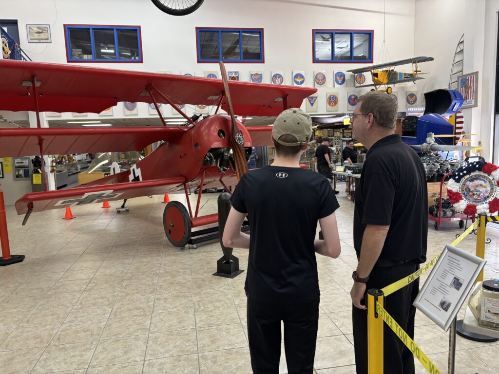 A red vintage biplane is displayed in a museum setting. Two people are standing in the foreground, looking at the plane. The room is filled with aviation memorabilia, including model planes and informational displays. The floor is tiled, and there are orange cones around the plane. The walls have various aviation-themed decorations and plaques.