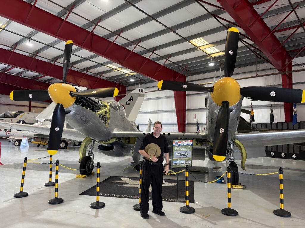 A man is standing in front of a vintage twin-engine aircraft inside a hangar. The aircraft has two large propellers with yellow tips and a silver body. The hangar has a high ceiling with red beams, and there are other aircraft visible in the background. The area is cordoned off with black and yellow stanchions.