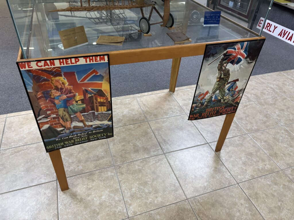 The image shows a display case with a model airplane inside, labeled with a sign that reads "Constructed and Donated by William J. Pritch." The case is supported by wooden legs, and two vintage posters are attached to the front. The left poster features a woman and child in front of a damaged building with a Union Jack flag, and the text "We Can Help Them! We Can Rush Relief to Britain." The right poster depicts a soldier holding a Union Jack flag, with the text "British War Relief Society." Both posters are from the British War Relief Society, located at 730 Fifth Avenue, New York. The floor is tiled, and there are other display items visible in the background.