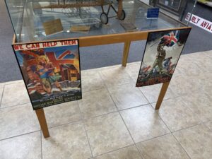 The image shows a display case with a model airplane inside, labeled with a sign that reads "Constructed and Donated by William J. Pritch." The case is supported by wooden legs, and two vintage posters are attached to the front. The left poster features a woman and child in front of a damaged building with a Union Jack flag, and the text "We Can Help Them! We Can Rush Relief to Britain." The right poster depicts a soldier holding a Union Jack flag, with the text "British War Relief Society." Both posters are from the British War Relief Society, located at 730 Fifth Avenue, New York. The floor is tiled, and there are other display items visible in the background.