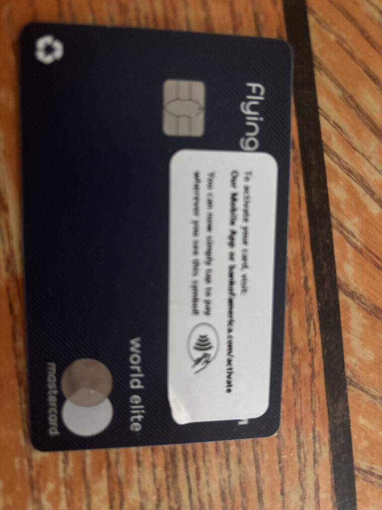 The image shows a dark-colored credit card with a chip on the left side. The card has the word "Flying" and "world elite" written on it, along with the Mastercard logo. There is a white sticker on the card with instructions for activation, including a website link and a contactless payment symbol. The card is placed on a wooden surface.