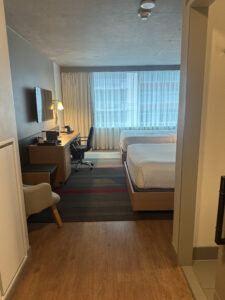 A modern hotel room with two neatly made beds on the right side. There is a large window with sheer curtains allowing natural light to enter. On the left, there is a desk with a chair, a lamp, and a wall-mounted TV above it. The floor is a combination of wood and a striped carpet. A light-colored armchair is positioned near the entrance.