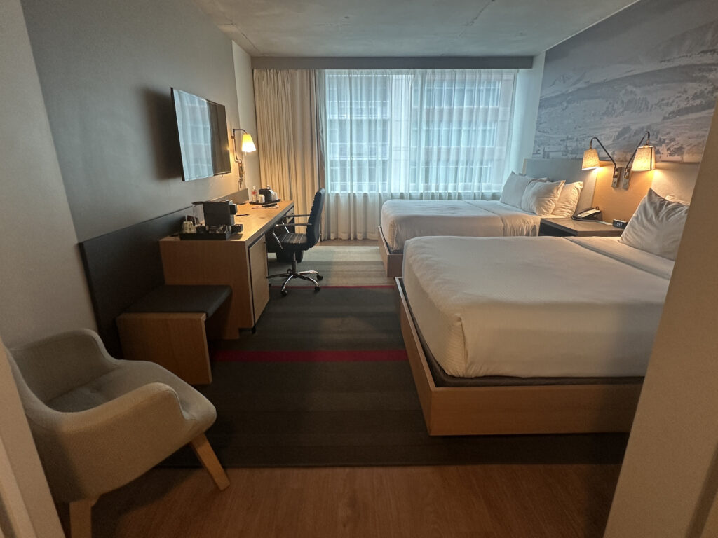 The image shows a hotel room with two neatly made double beds. There is a large window with sheer curtains allowing natural light to enter. A desk with a chair is positioned against the wall, featuring a coffee maker and a lamp. A flat-screen TV is mounted on the wall above the desk. An armchair is placed near the entrance. The room has a modern and clean design.