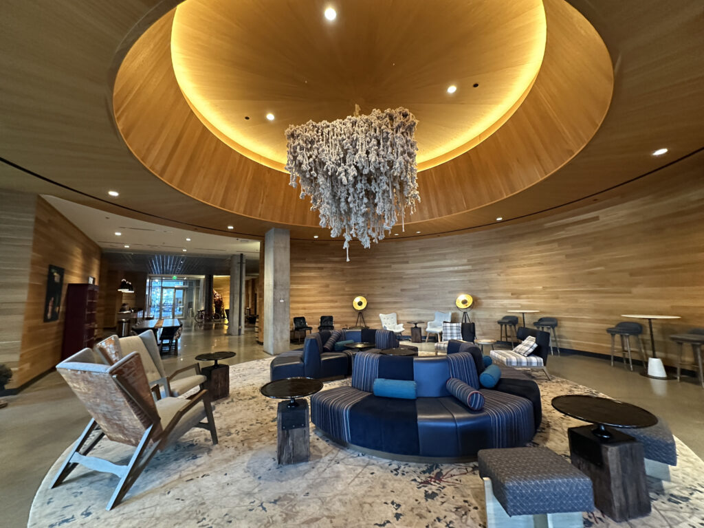 The image shows a modern, stylish hotel lobby with a circular seating arrangement in the center. The seating is upholstered in dark blue and striped fabric, surrounded by small tables and additional chairs. The ceiling features a large, circular wooden design with a unique, textured chandelier hanging from the center. The walls are lined with wood paneling, and there are bar stools along the right side. The overall ambiance is warm and inviting, with soft lighting and contemporary decor.