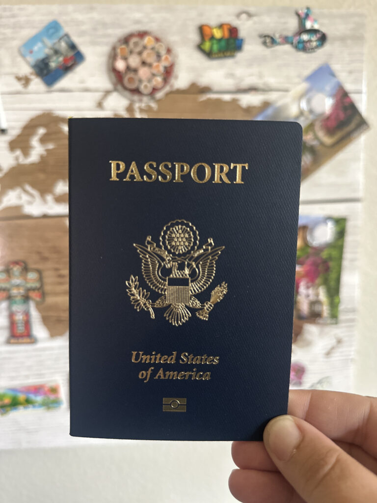 A person is holding a United States passport in front of a world map. The passport is dark blue with gold lettering and the U.S. emblem. The background features various colorful magnets and a blurred map design.
