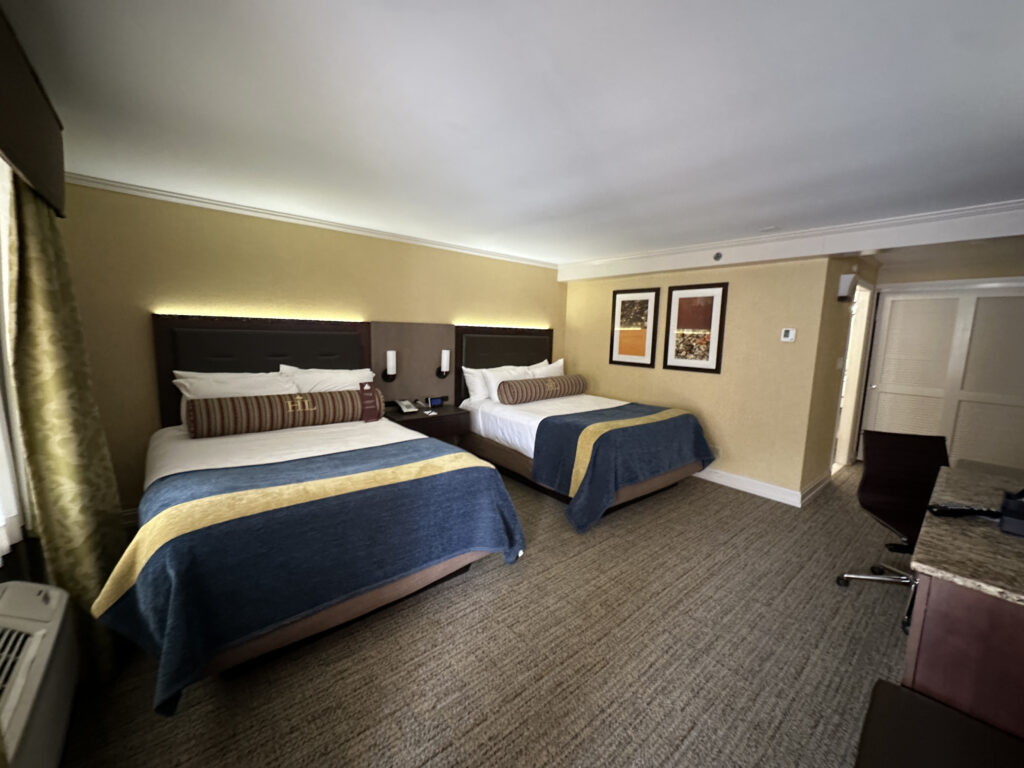 The image shows a hotel room with two double beds. Each bed has a dark headboard, white sheets, and a decorative bolster pillow with a striped pattern. The beds are covered with blue and yellow bedspreads. Above the beds, there are two framed artworks on the wall. The room has beige walls and carpeted flooring. To the right, there is a desk with a chair and a granite countertop. A window with curtains is partially visible on the left side of the image.