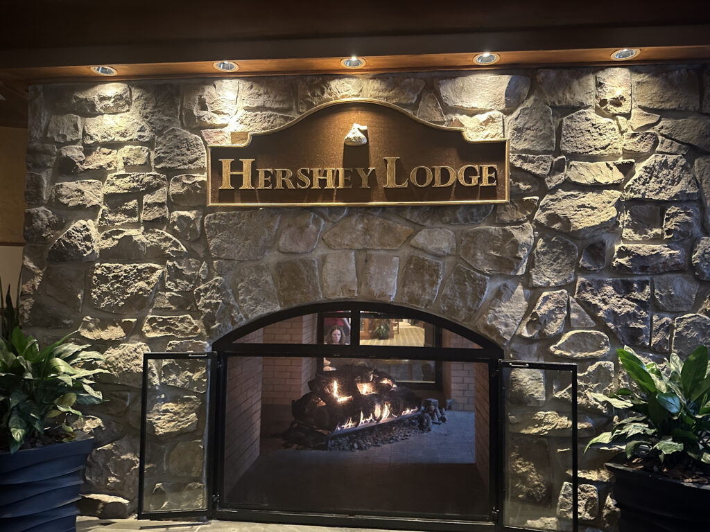 The image shows a stone fireplace with a lit fire inside. Above the fireplace is a wooden sign with the text "Hershey Lodge." The area is well-lit with spotlights, and there are potted plants on either side of the fireplace.