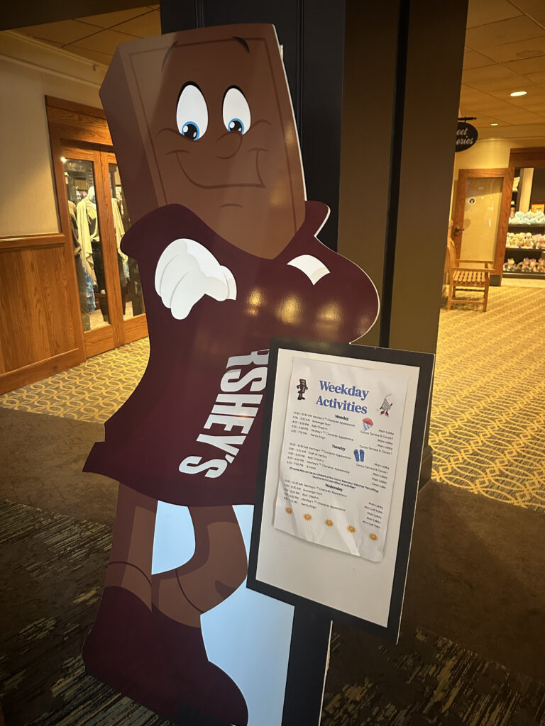 The image shows a large, cartoonish cutout of a smiling chocolate bar character wearing gloves and shoes, with the word "HERSHEY'S" on its body. Next to it is a sign displaying "Weekday Activities" with a list of events and times. The setting appears to be indoors, with a carpeted floor and wooden accents in the background.