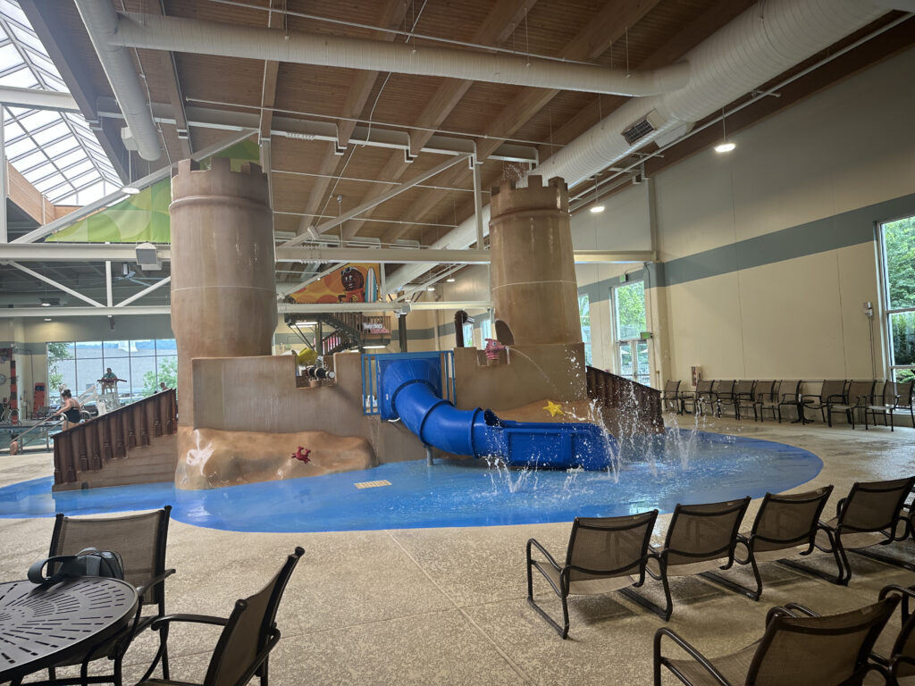 The image shows an indoor water play area designed like a castle. It features two large castle towers connected by walls, with a blue water slide emerging from one side. Water is splashing around the slide's exit into a shallow pool. The area is surrounded by seating, including lounge chairs and a table with chairs. The ceiling has exposed beams and ductwork, and large windows provide natural light.