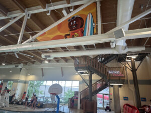 The image shows an indoor swimming pool area with a high ceiling and wooden beams. There are people near the pool, including a lifeguard on a chair and several swimmers. A basketball hoop is positioned near the pool. Above, there is a large banner featuring a cartoon character holding a surfboard. A staircase with a sign that reads "Twizzlers twists" is visible in the background. The area is well-lit with large windows allowing natural light to enter.