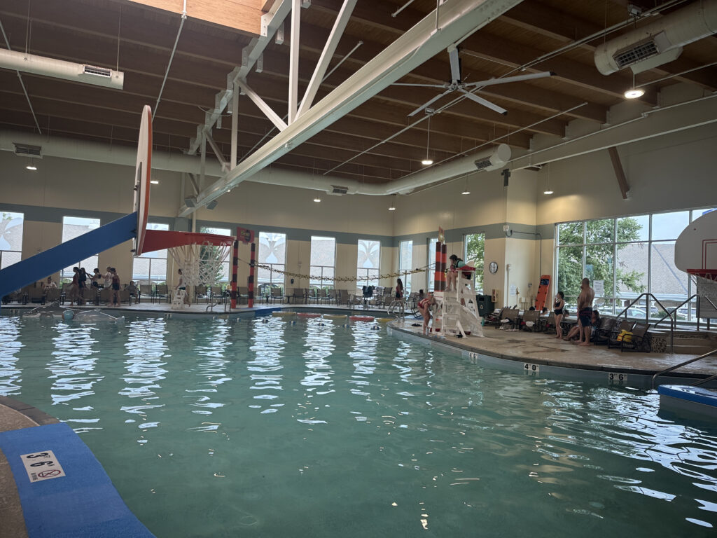 The image shows an indoor swimming pool with a basketball hoop and a slide. There are people around the pool, including a lifeguard sitting on a high chair. The pool area has large windows allowing natural light to enter, and the ceiling has exposed beams and lighting fixtures. The water is calm, and there are some poolside chairs and equipment visible.