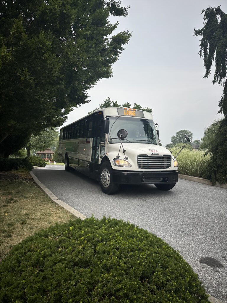 A white bus with a sign displaying "Lodge" on the front is driving on a paved road. The road is surrounded by lush greenery, including trees and bushes. The sky is overcast.