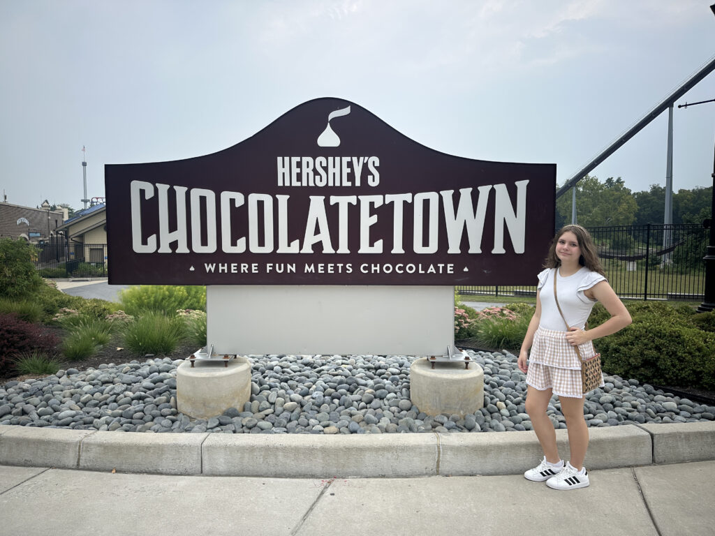 A person is standing next to a large sign that reads "Hershey's Chocolatetown: Where Fun Meets Chocolate." The sign is surrounded by landscaping with rocks and greenery. The person is wearing a white top, a plaid skirt, white sneakers, and is carrying a small bag. The background includes trees and part of a building.