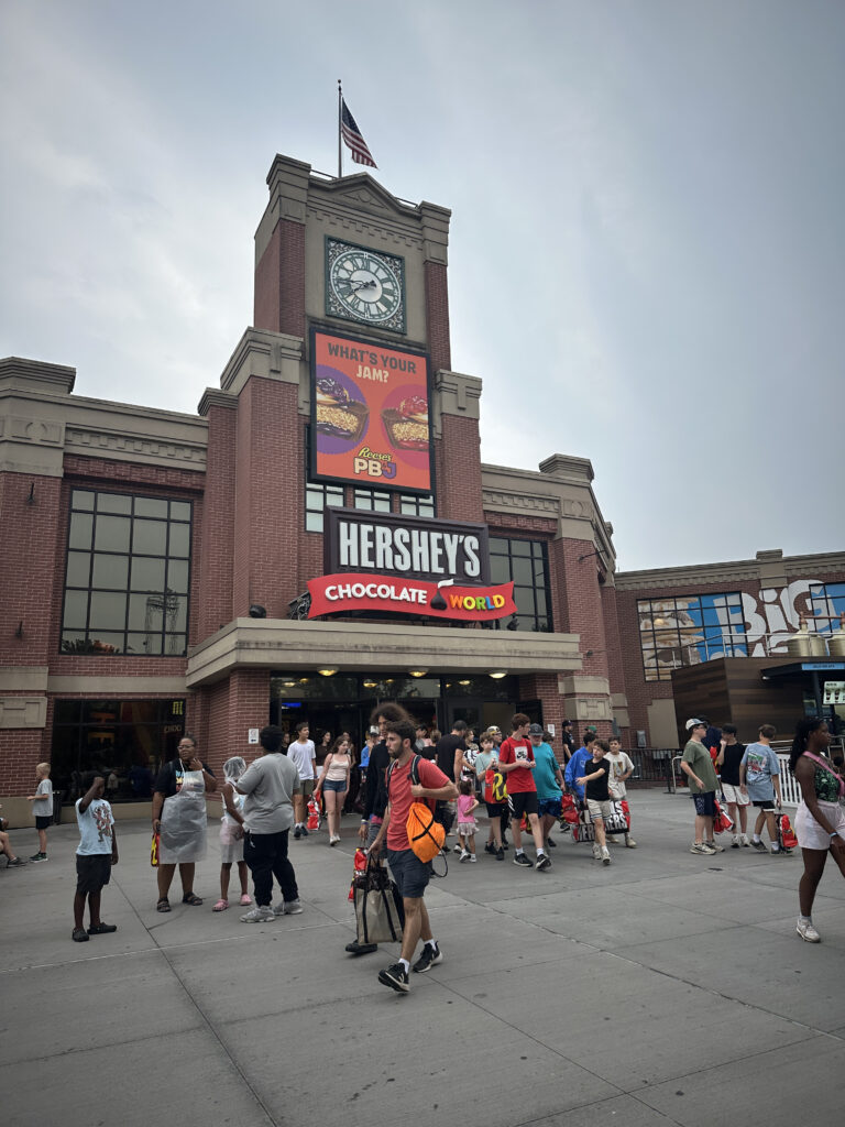 The image shows the entrance to Hershey's Chocolate World, a large brick building with a clock tower and an American flag on top. There is a sign above the entrance that reads "Hershey's Chocolate World." A digital billboard displays an advertisement for Reese's PB&J with the text "What's Your Jam?" In front of the building, a crowd of people, including families and individuals, are walking and standing around, some carrying bags. The sky is overcast.