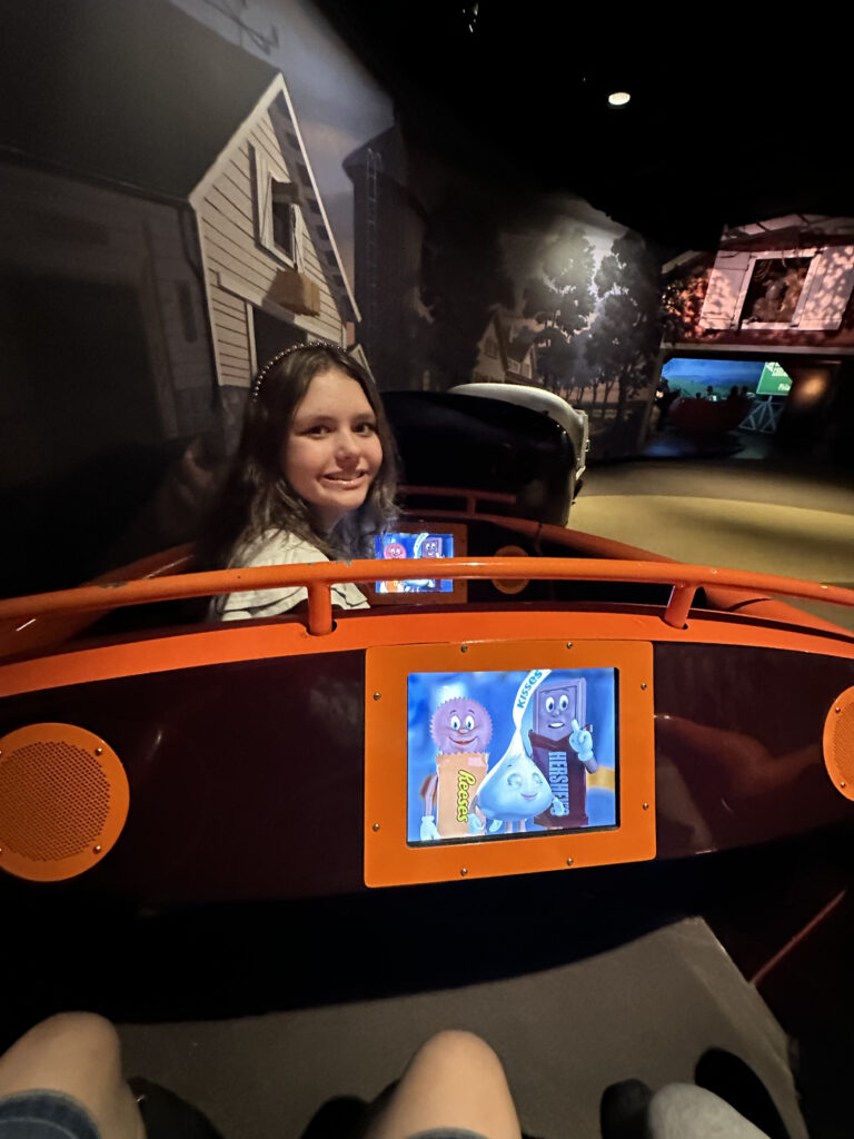 A person is sitting in a theme park ride vehicle, smiling at the camera. The vehicle has an orange and maroon color scheme with a screen displaying animated characters from Reese's, Hershey's, and Kisses. The background features a mural of a house and trees, creating an immersive environment.
