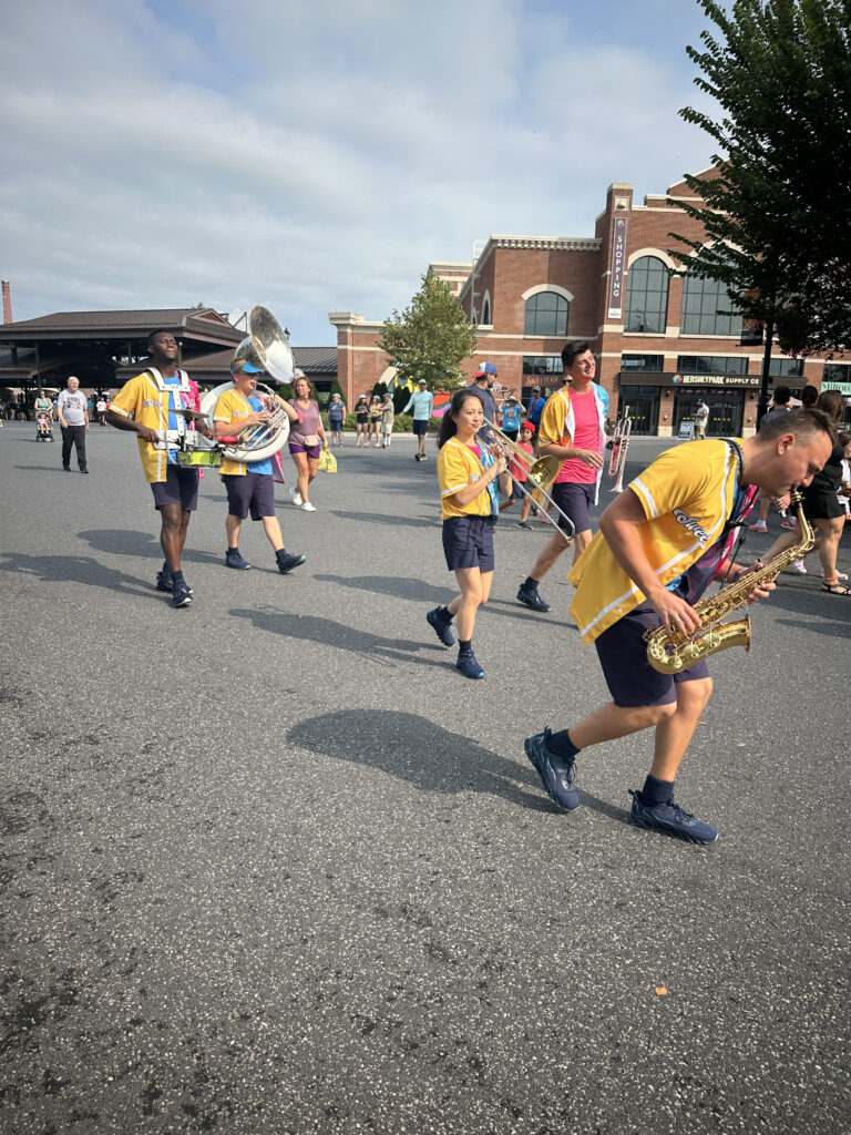 A marching band is performing in an outdoor setting. The band members are wearing yellow and blue uniforms and playing various instruments, including a saxophone, trombone, trumpet, and sousaphone. They are marching on a paved surface with a brick building and trees in the background. People are watching the parade from the sides.