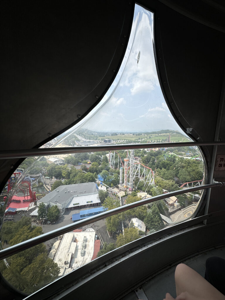 View from inside a Ferris wheel cabin, looking out through a teardrop-shaped window. The scene outside shows an amusement park with roller coasters, trees, and various buildings. The sky is partly cloudy, and the landscape extends into the distance.