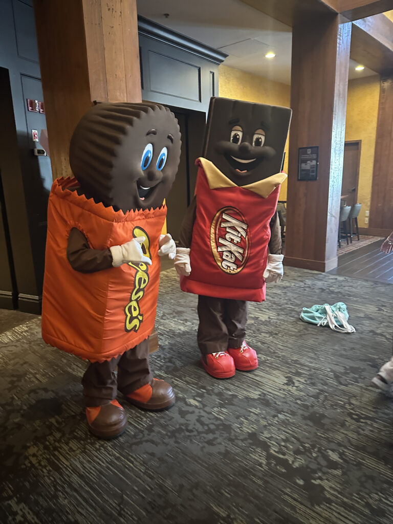 The image shows two people dressed in large, colorful costumes. One is wearing a Reese's Peanut Butter Cup costume, which is orange with a round, textured top resembling the candy. The other is in a Kit Kat bar costume, which is red with a rectangular shape. Both costumes have cartoonish facial features and are standing indoors on a carpeted floor.