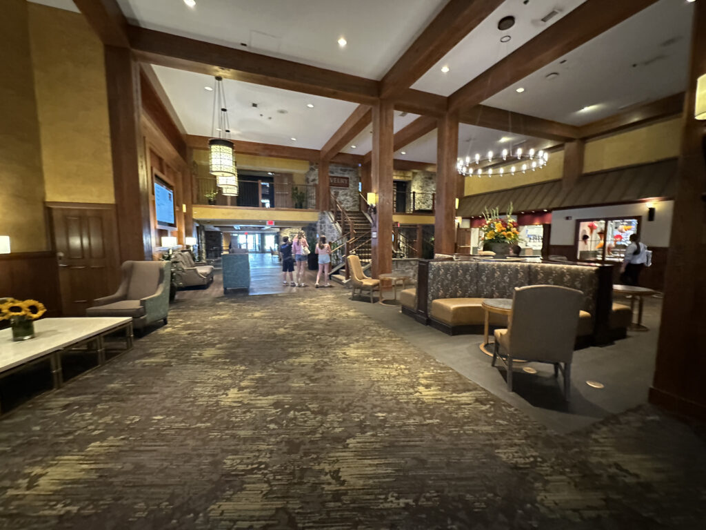 The image shows the interior of a spacious hotel lobby. The area features a high ceiling with wooden beams and modern lighting fixtures. There are several seating areas with cushioned chairs and sofas. A large floral arrangement is placed on a table in the center. In the background, a staircase leads to an upper level, and a few people are standing near the entrance. The decor is warm and inviting, with a mix of wood and stone elements.