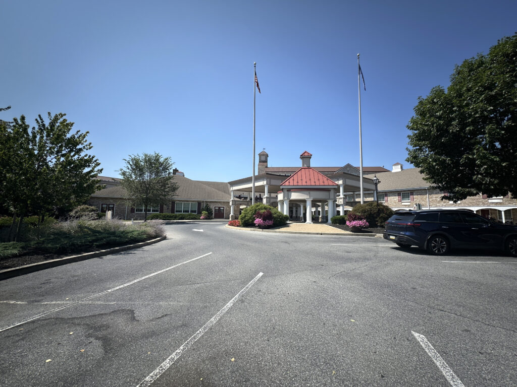 The image shows the exterior of a large building with a red-roofed entrance and two flagpoles in front. The building is surrounded by a parking area with a few cars and landscaped with trees and bushes. The sky is clear and blue.