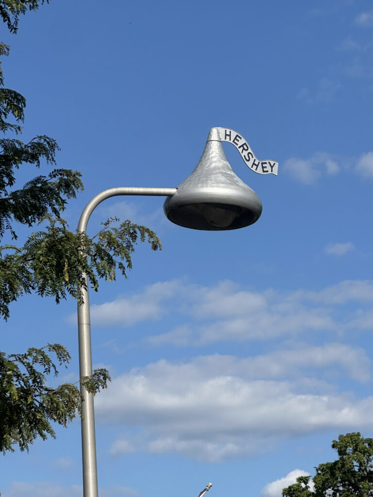 A streetlight designed to resemble a Hershey's Kiss, complete with a silver wrapper and a small flag that reads "HERSHEY," set against a blue sky with some clouds and tree branches in the foreground.