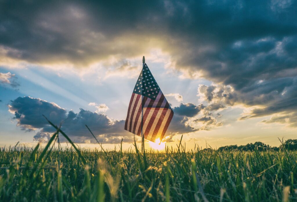 A small American flag is planted in a grassy field, with the sun setting in the background. The sky is partly cloudy, and the sunlight creates a dramatic effect with rays shining through the clouds.