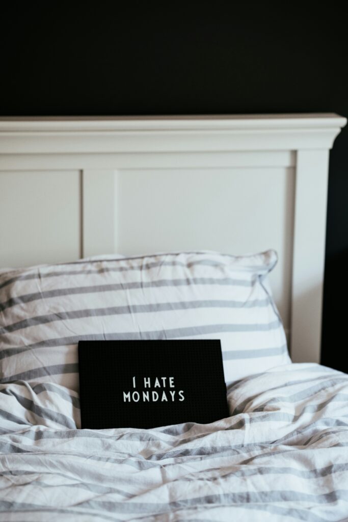 A bed with a white and gray striped duvet and pillows. A black letter board is placed on the bed with the text "I HATE MONDAYS." The headboard is white, and the background wall is dark.