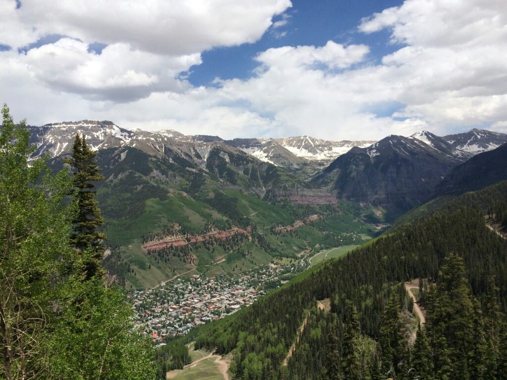 A scenic view of a mountainous landscape with snow-capped peaks under a partly cloudy sky. In the foreground, there are lush green trees and a small town nestled in a valley. The town is surrounded by dense forests and rolling hills.