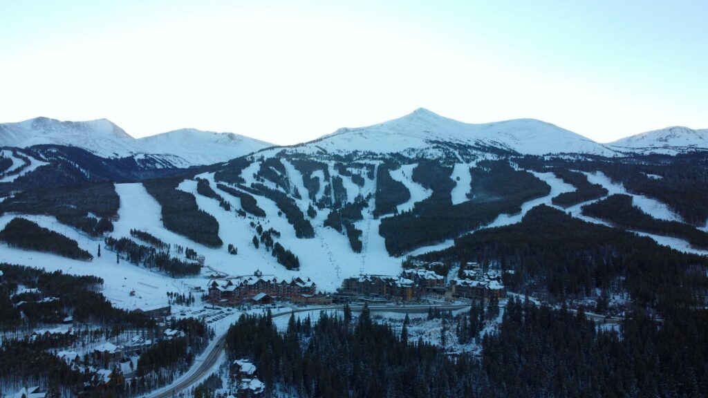 Aerial view of a snow-covered mountain ski resort. The image shows multiple ski trails winding down the slopes, surrounded by dense forests. At the base of the mountain, there are buildings and lodges, with a road leading through the area. The sky is clear, and the scene is set in a winter landscape.
