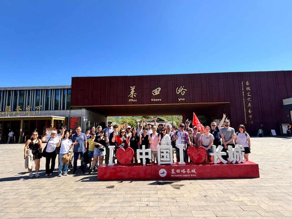 A group of people is posing for a photo in front of a building with Chinese characters on it. They are standing behind a red sign that reads "I ❤️ 中国 I ❤️ 长城" with "Mutianyu Great Wall" written below. The sky is clear and blue.