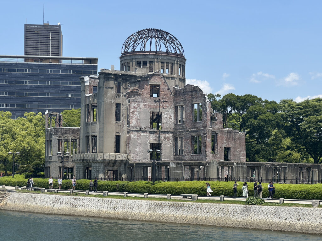 The image shows the Hiroshima Peace Memorial, also known as the Atomic Bomb Dome, a partially preserved building with a skeletal dome structure on top. It is surrounded by greenery and a river in the foreground. People are walking along a path near the building. A modern building is visible in the background under a clear blue sky.