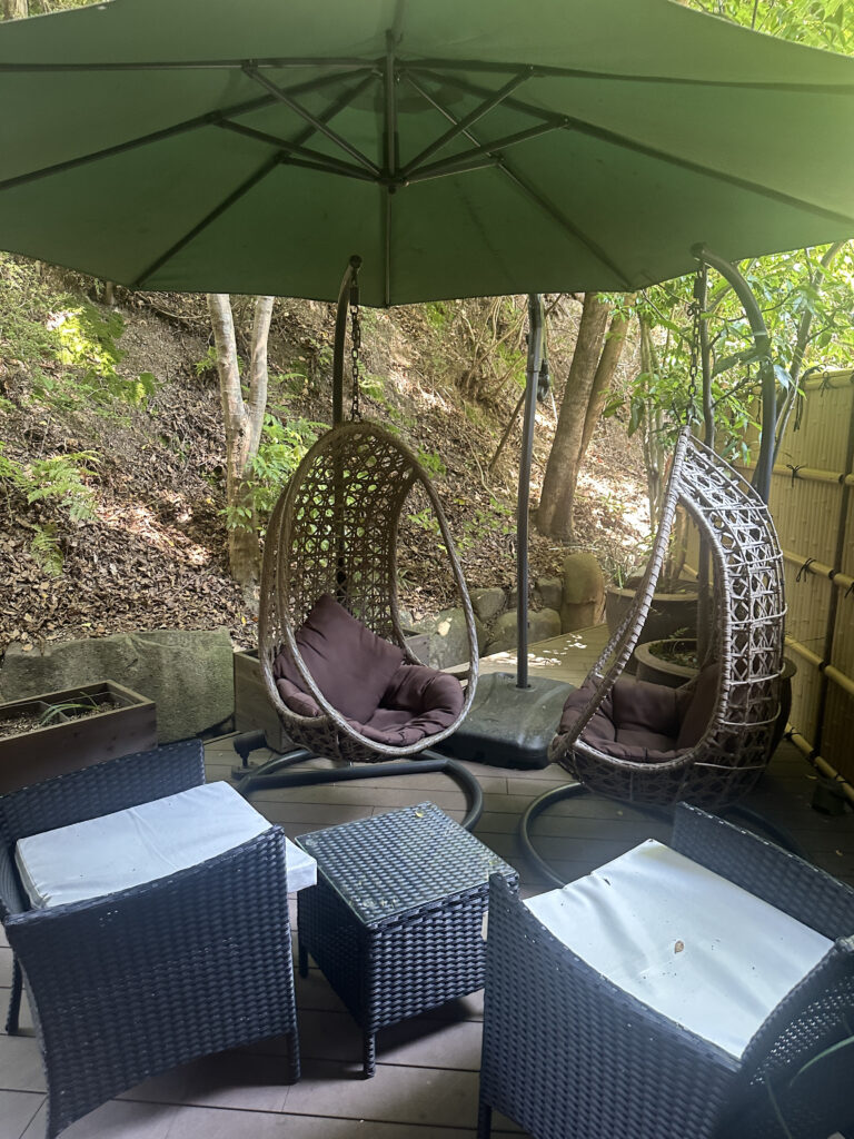 The image shows an outdoor seating area with two hanging wicker egg chairs, each with a cushion inside. There is a large green umbrella providing shade. In front of the chairs, there are two small wicker tables and two wicker chairs with white cushions. The area is surrounded by trees and greenery, creating a natural and relaxing atmosphere.