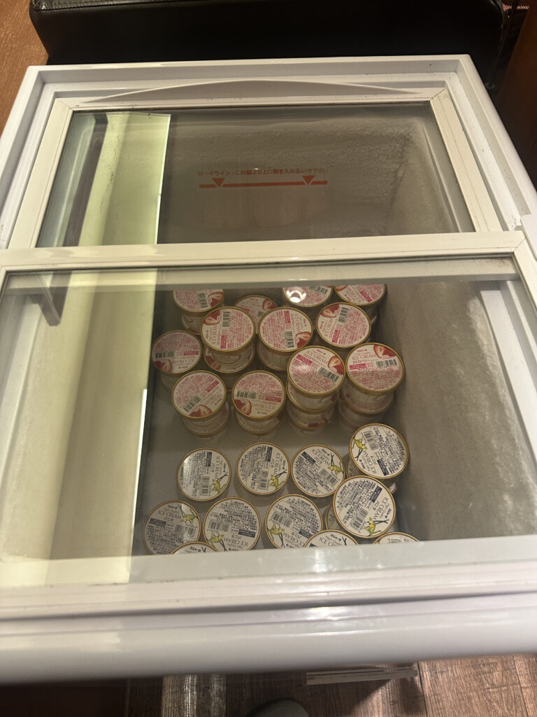 The image shows the interior of a freezer with a glass lid, containing several small containers of ice cream. The containers have labels with text and images, and they are stacked neatly inside the freezer. The interior walls of the freezer have a frosty appearance.