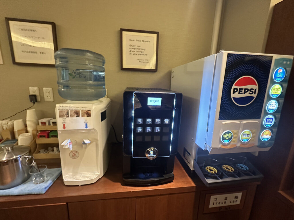 The image shows a beverage station with three machines. On the left is a water dispenser with a large water jug on top, labeled "Aqua Clara," offering hot and cold water options. In the middle is a coffee machine with various selection buttons. On the right is a Pepsi-branded soda fountain with multiple drink options. Below the machines, there are trays for cups and a trash can labeled in both Japanese and English.