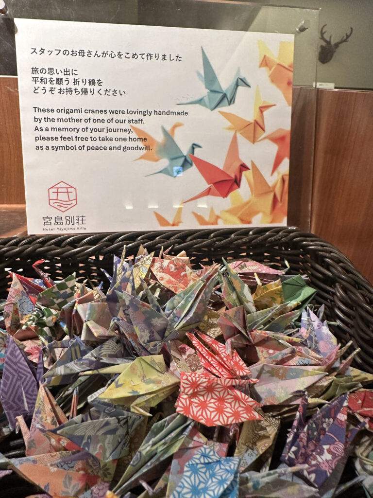 A basket filled with colorful origami cranes is displayed in the foreground. Above the basket, there is a sign with text in both Japanese and English. The English text explains that the origami cranes were handmade by the mother of a staff member and invites visitors to take one home as a symbol of peace and goodwill. The background features an image of more origami cranes.