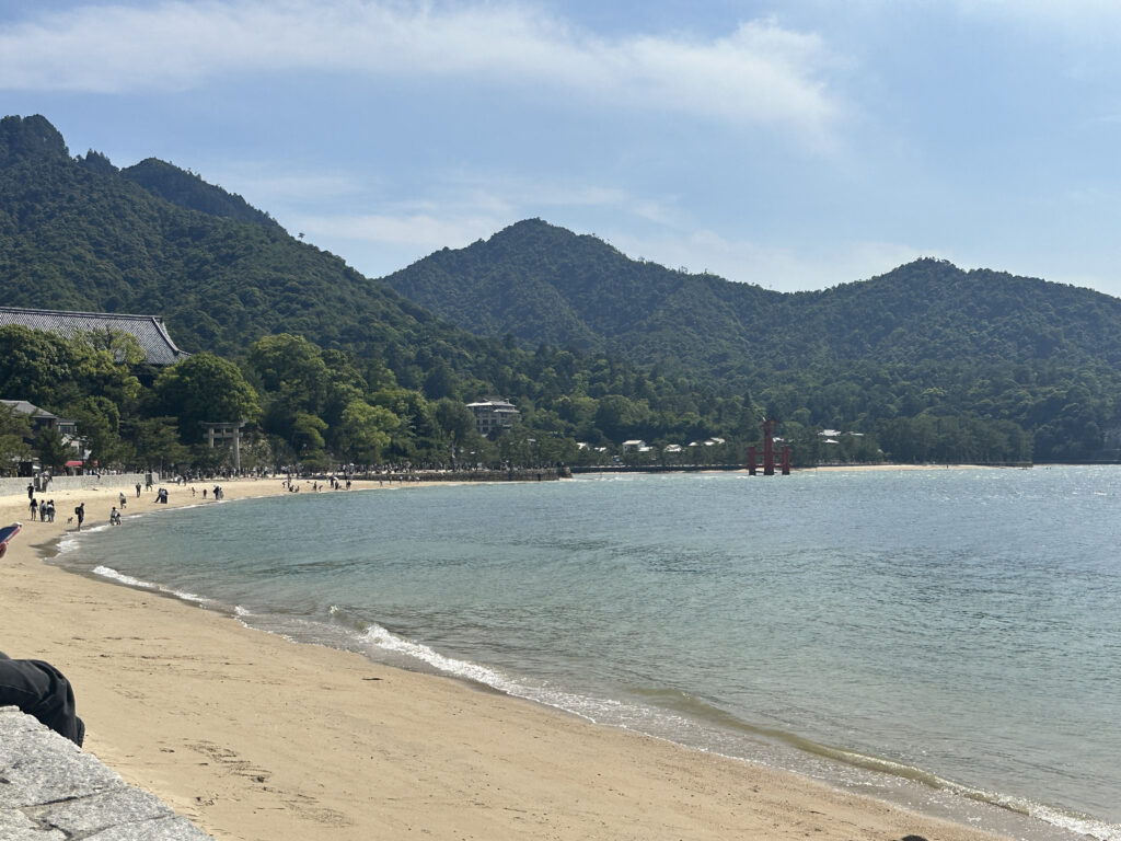 A scenic view of a sandy beach with gentle waves lapping at the shore. In the background, lush green mountains rise under a partly cloudy sky. A red torii gate stands in the water near the shoreline, and people are walking along the beach and near the trees. A traditional building with a tiled roof is partially visible on the left.