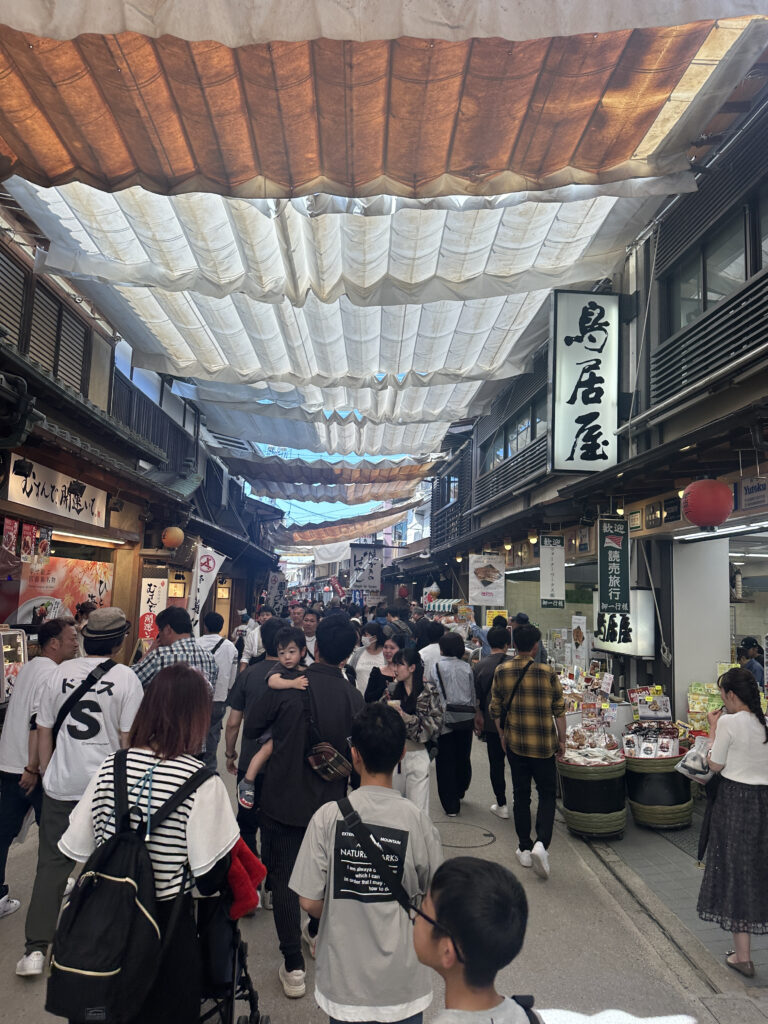 A bustling outdoor market street with many people walking in both directions. The street is covered with fabric canopies providing shade. Shops line both sides, displaying various goods and signs with Japanese characters. The atmosphere is lively and crowded.