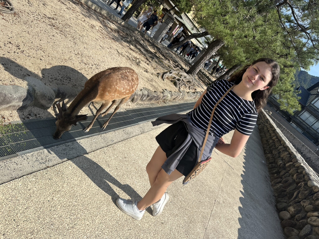 A young woman is standing on a paved path, smiling at the camera. She is wearing a black and white striped shirt, black shorts, and white sneakers, with a sweater tied around her waist and a crossbody bag. Next to her, a deer is grazing near a metal grate. The background features trees, a stone wall, and a few people walking in the distance.