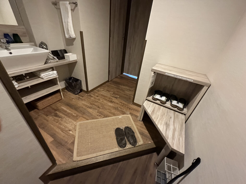 The image shows a small entryway area with wooden flooring. There is a mat on the floor with a pair of black slippers on it. To the right, there is a wooden shelf with two pairs of white slippers. On the left, there is a sink with a towel hanging above it, and shelves underneath containing more towels and a basket. A trash bin is placed next to the sink.
