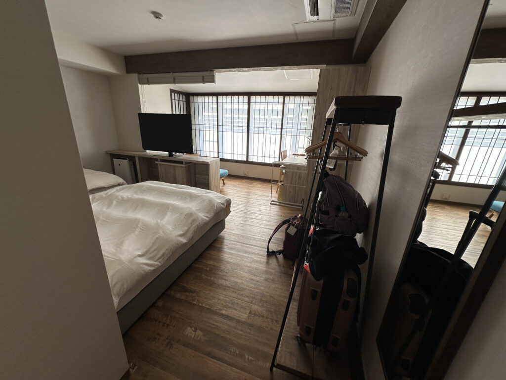 The image shows a modern hotel room with wooden flooring. There is a bed with white bedding on the left side. A flat-screen TV is placed on a wooden stand in front of the bed. Large windows with grid-like frames allow natural light to enter the room. On the right, there is a luggage rack with hangers, a suitcase, and a backpack. A full-length mirror is also visible on the right wall.