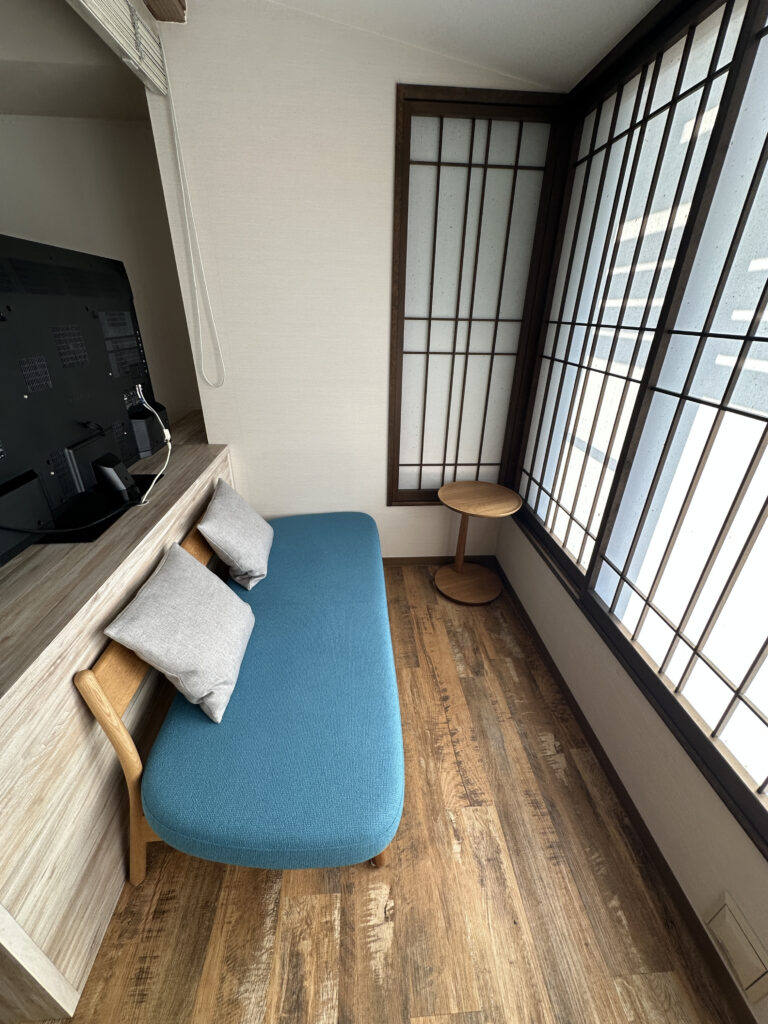 The image shows a small, minimalist room with a wooden floor. There is a blue cushioned bench with two gray pillows against a wooden partition. A flat-screen TV is mounted on the partition. To the right, there is a large window with a grid pattern, and a small round wooden side table is placed near the window. The room has a clean and simple design.